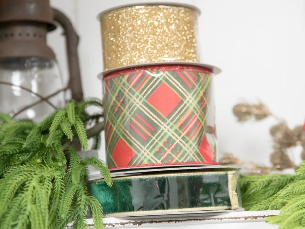 Two spools of ribbon, one gold and one plaid pattern, on a surface with greenery.